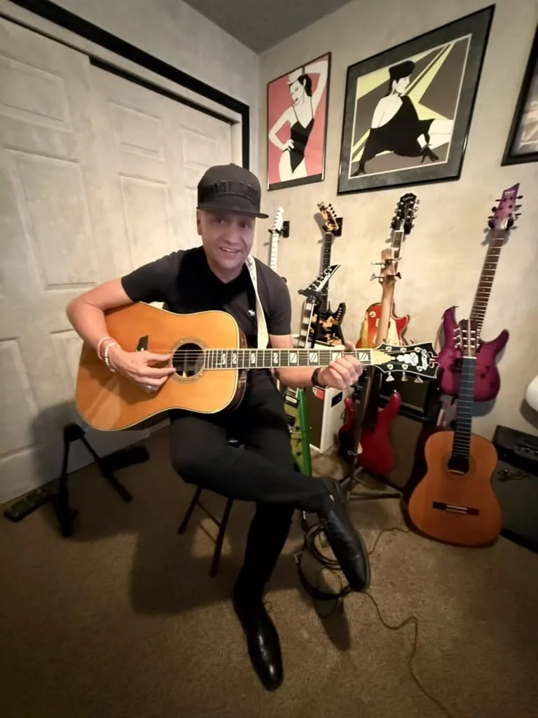 A guitarist in a black cap sits with an acoustic guitar in front of a wall of electric guitars